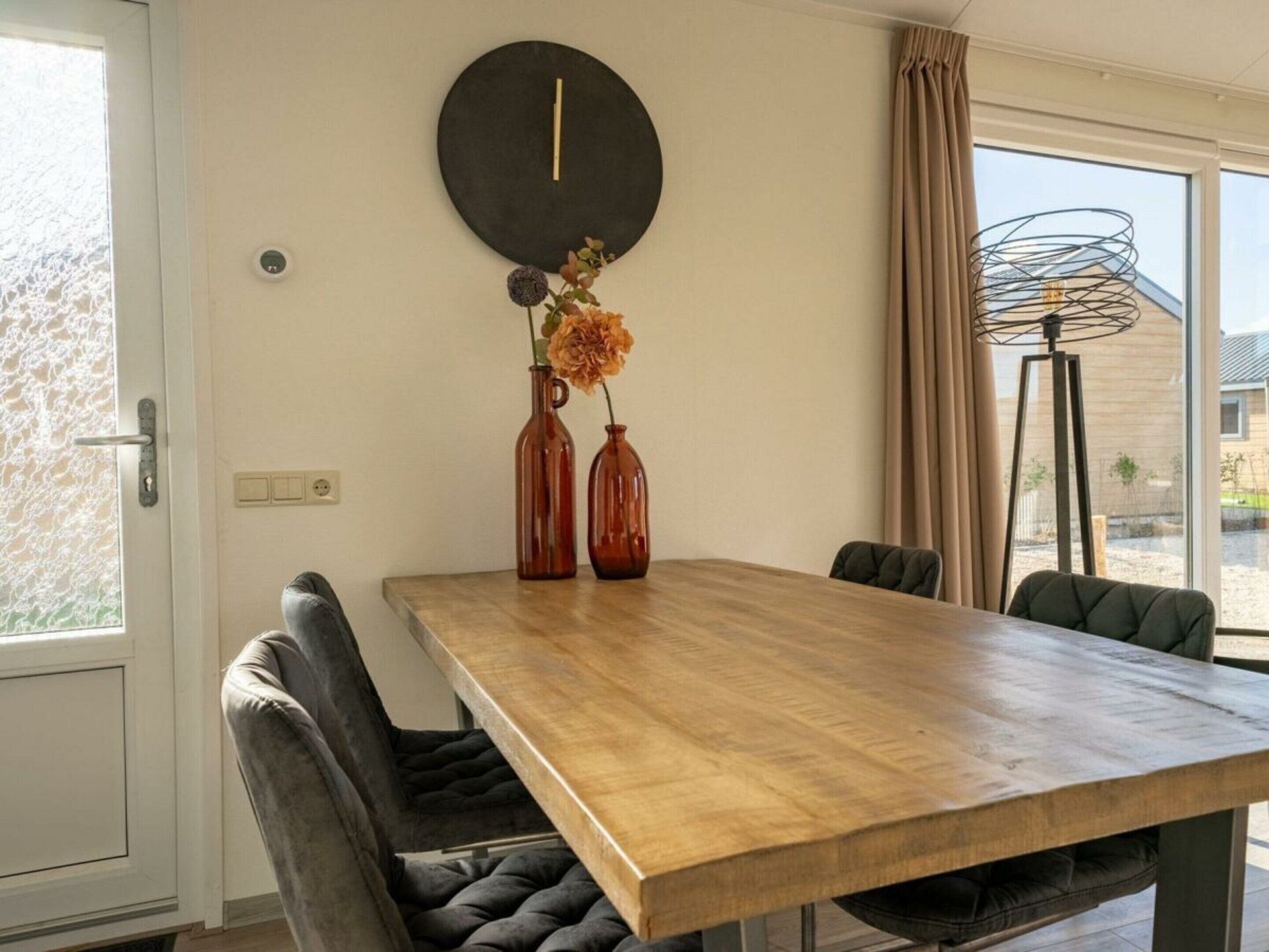 A dining area with a wooden table, two chairs, a wall clock, and a vase with flowers.