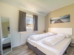 Ein Schlafzimmer mit einem Bett, einem Fenster mit Vorhängen, einem Heizkörper, einem Spiegel und einem Bild an der Wand.