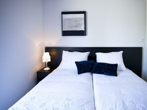 A neatly made bed with white bedding and two dark blue pillows, a bedside lamp, and a framed picture on the wall. - Holiday Home in Velsen-zuid With Terrace (Velsen-Zuid)