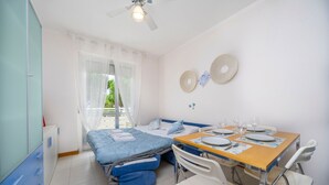 Une pièce de taille modeste comprenant un lit, une petite table à manger avec des chaises, un ventilateur de plafond et une fenêtre avec des rideaux.