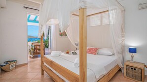 Ein Bett mit Baldachin, ein Nachttisch mit Lampe und Blick auf das Meer durch ein Fenster.