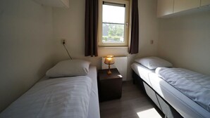 Alojamiento pequeño de una sola habitación con dos camas individuales, una ventana con cortinas, una mesita de noche con lámpara y un radiador.