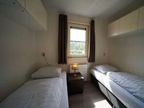 Een kleine, eenvoudige kamer met twee éénpersoonsbedden, een raam met gordijnen, een nachtkastje met lamp en een radiator.