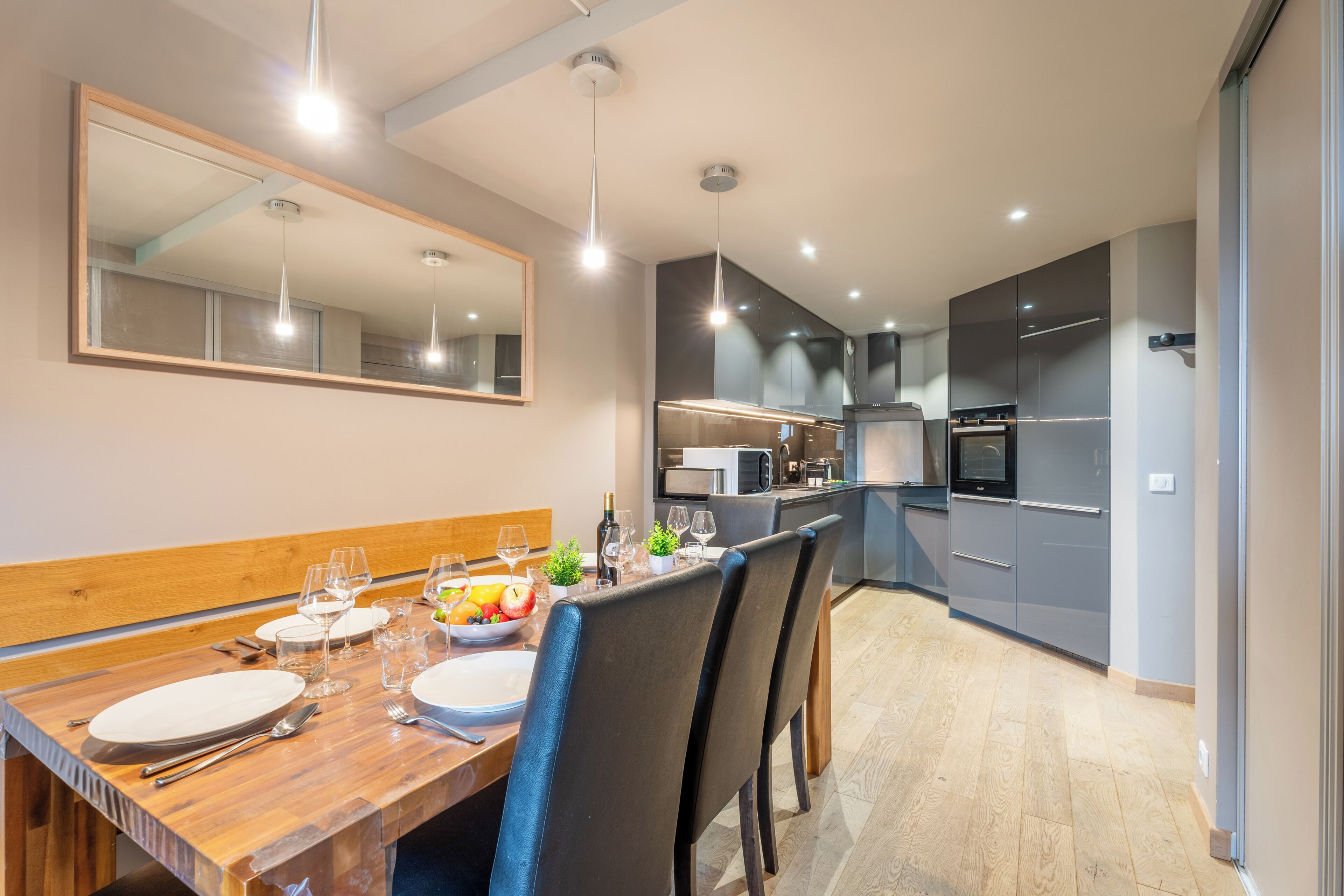 A modern dining area with a wooden table set for four, a large mirror, and a kitchen in the background.