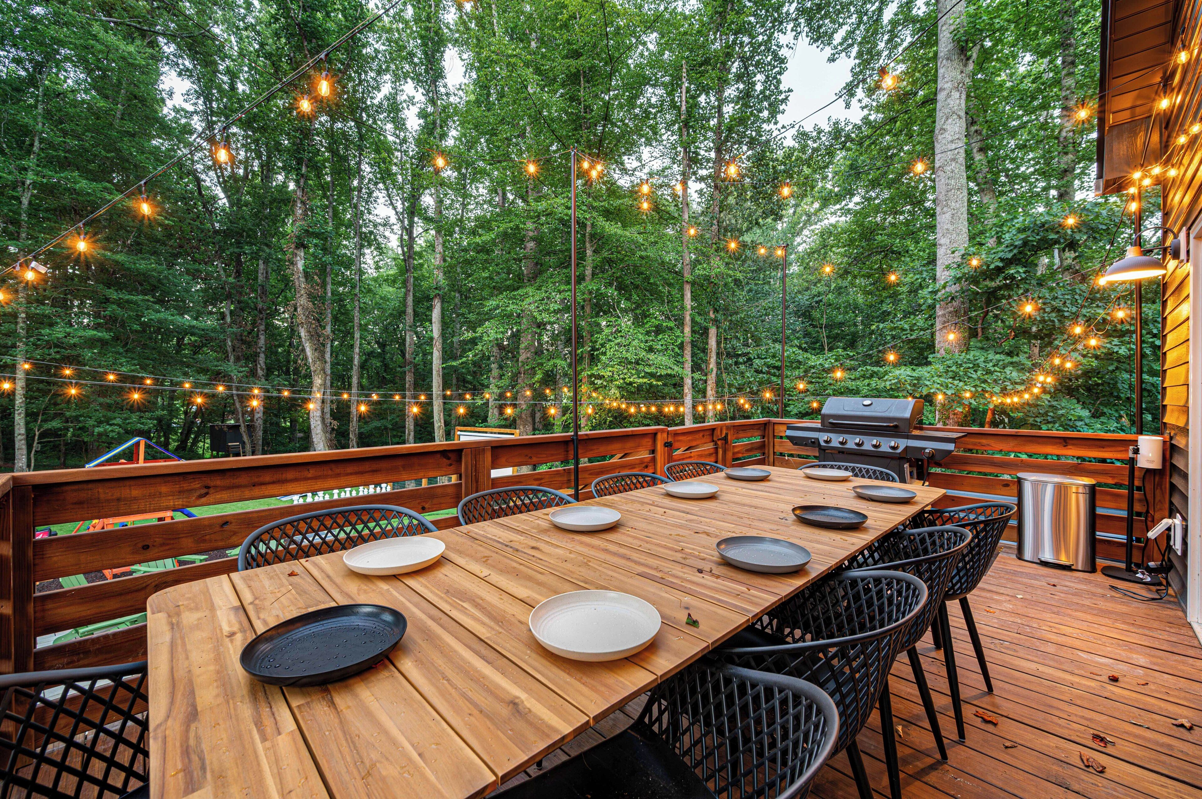 An outdoor deck with a wooden table set for dining, surrounded by chairs, and string lights.