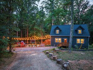 Une cabane dotée d’une terrasse, entourée d’arbres, et illuminée de guirlandes lumineuses.