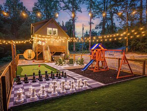 Une cabane en bois avec une terrasse, une balançoire et un grand jeu d’échecs.