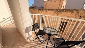 Un balcon avec une table ronde en verre et deux chaises, une balustrade blanche et une vue sur les bâtiments en arrière-plan.