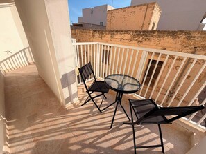 Ein Balkon mit rundem Glastisch und zwei Stühlen, weißem Geländer und Blick auf Gebäude im Hintergrund.