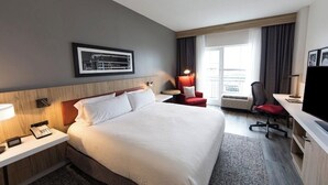 Ein Hotelzimmer mit Bett, Nachttisch mit Telefon, Schreibtisch mit Stuhl, Fernseher, Fenster mit Vorhängen und einem gerahmten Bild an der Wand.