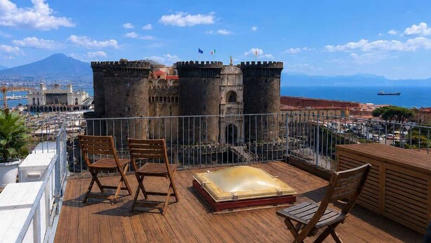 Um deck de madeira com área de estar externa e banheira de hidromassagem, com vista para um castelo histórico e o mar.