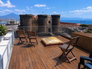 Eine Holzterrasse mit Sitzgelegenheiten im Freien und einem Whirlpool, mit Blick auf eine historische Burg und das Meer.