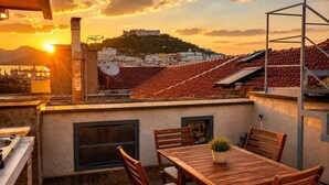 Terraço no telhado com mesa e cadeiras, uma pequena planta em vaso, e vista para uma colina com edifícios.