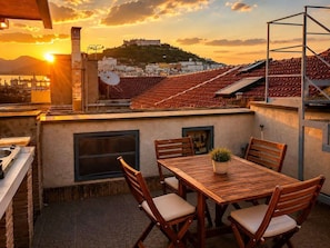Eine Dachterrasse mit Tisch und Stühlen, einem kleinen Topfpflanzen und Blick auf einen Hügel mit Gebäuden.