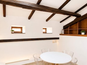 Ein Zimmer mit weißem Tisch und Stühlen, Holzbalken an der Decke und kleinen Fenstern mit Pflanzen.