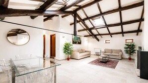 Un vaste espace de vie doté d’un plafond haut, de poutres en bois, d’une table à manger en verre, de deux canapés et de plantes en pot.