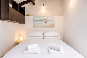 Ein ordentlich bezogenes Bett mit weißen Leinen und Handtüchern, ein Gemälde einer Küstenlandschaft darüber und eine Nachttischlampe.