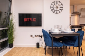 Ein moderner Essbereich mit einem Flachbildfernseher, der 'NETFLIX' anzeigt, einer Wanduhr und einem mit Tellern und Gläsern gedeckten Esstisch.