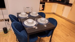 Ett matbord dukat med vita tallrikar, silverbestick och vinglas, omgivet av blå stolar i ett modernt kök med trägolv.