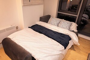 A neatly made bed with a dark headboard, a window with a view of the outside, and a radiator on the wall. - Luxury 2Bed 2Bath City Centre Apt | Balcony View (Leeds)
