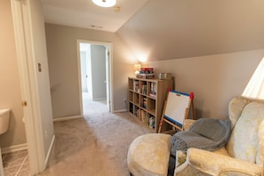 Una habitación con piso alfombrado, una puerta blanca, una estantería, una lámpara y una silla.