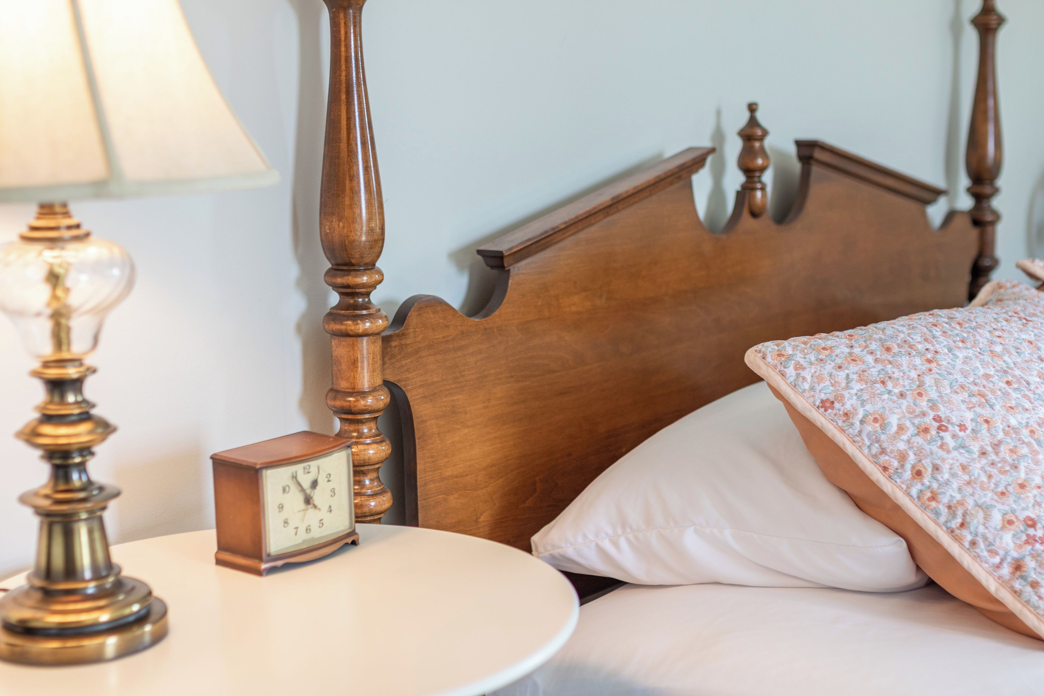 Uma cama com travesseiro estampado em floral, cabeceira de madeira e mesa de cabeceira com abajur e relógio.