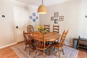 A dining area with a round wooden table surrounded by six wooden chairs, a woven pendant light, and a patterned rug.