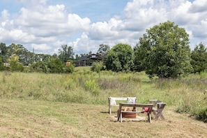An outdoor seating area with a rustic wooden bench and a table, surrounded by a grassy field and trees.
