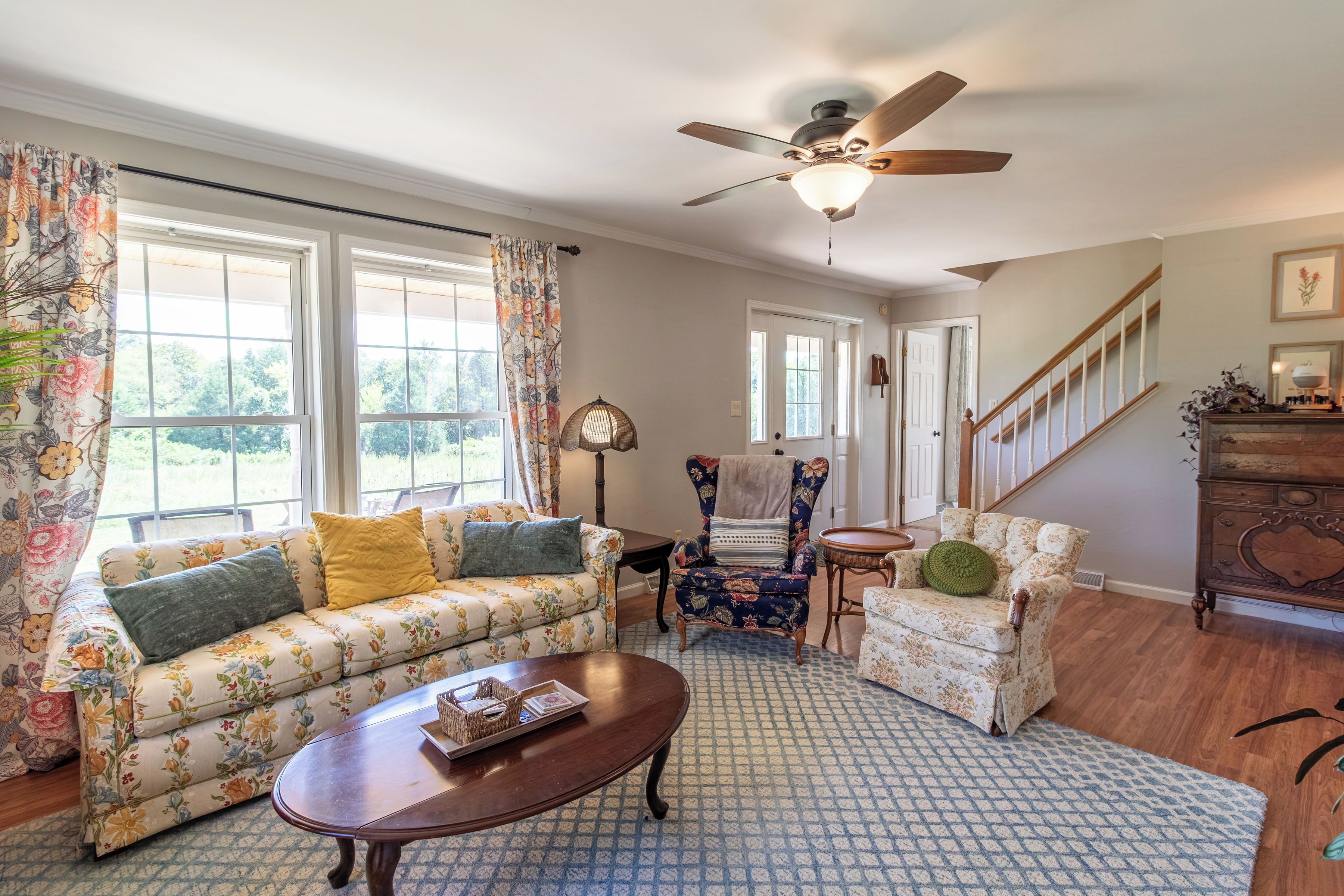 A living room with a floral sofa, a wooden coffee table, a patterned rug, a ceiling fan, and a staircase.