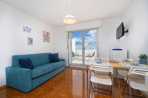 A living room with a teal sofa, a dining table with chairs, a TV mounted on the wall, and a sliding glass door leading to an outdoor area. - A comfortable 53-square-meter apartment, perfect for four people, located on the third floor (Scala A) of a building with an elevator (NOT suitable for people with disabilities) directly facing the seafront.The property is located in Lignano Sabbiadoro i (Lignano Sabbiadoro)