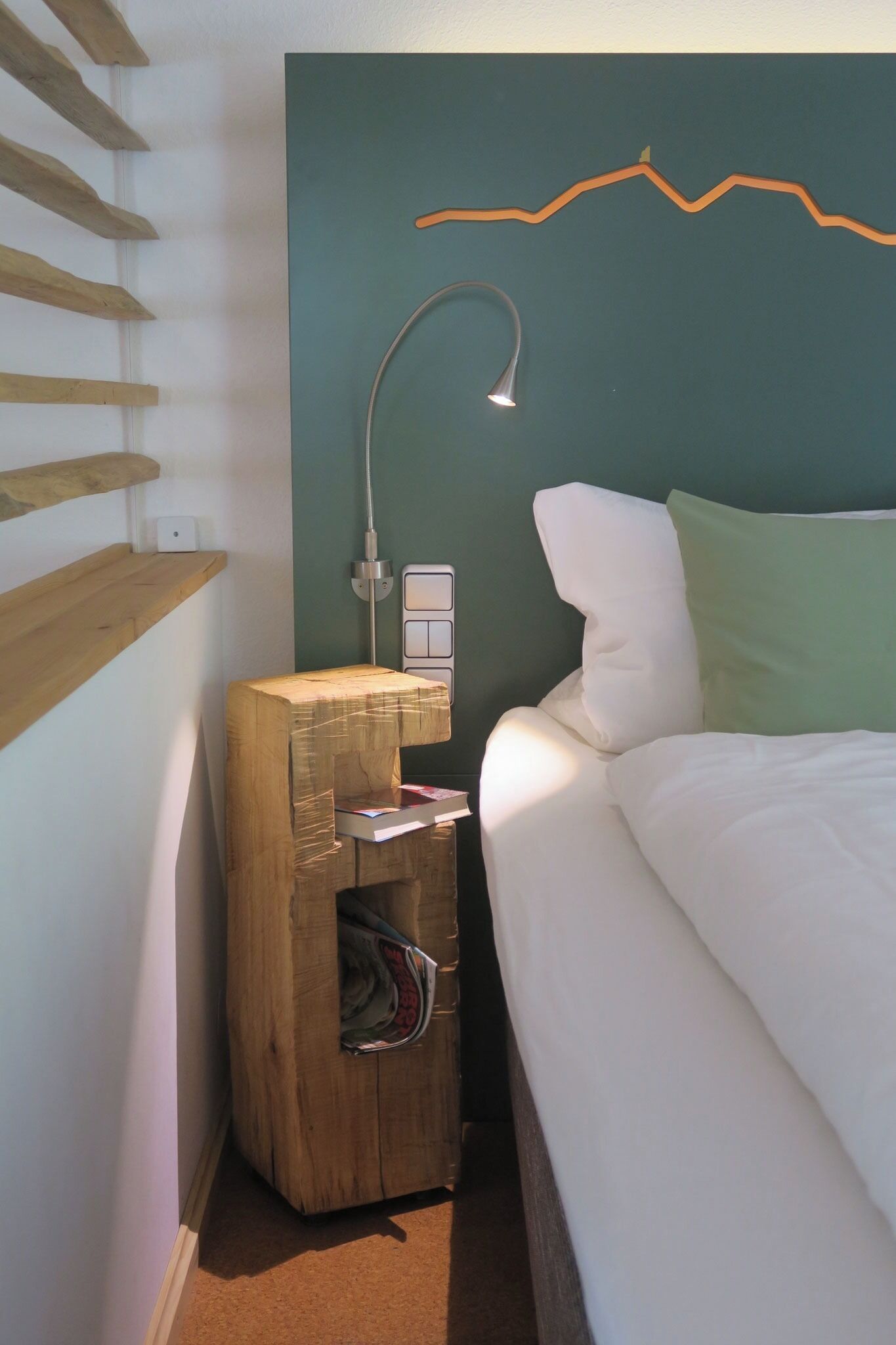 A bedroom with a bed, a wooden nightstand, a wall-mounted lamp, and a simple wall decoration.