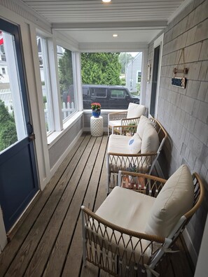 Eine überdachte Veranda mit Korbstühlen und einem kleinen Tisch, eine blaue Tür sowie Blick auf eine Straße und Grünflächen.