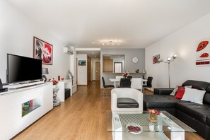 Ein modernes Wohnzimmer mit Flachbildfernseher, Glastisch, schwarzem Ledersofa und Essbereich mit weißem Tisch und Stühlen.