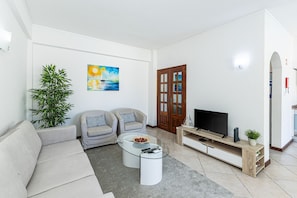 Ein Wohnzimmer mit einer Couch, zwei Sesseln, einem Couchtisch, einem Fernseher auf einem Holzboden, einem Bild an der Wand und einer Topfpflanze.