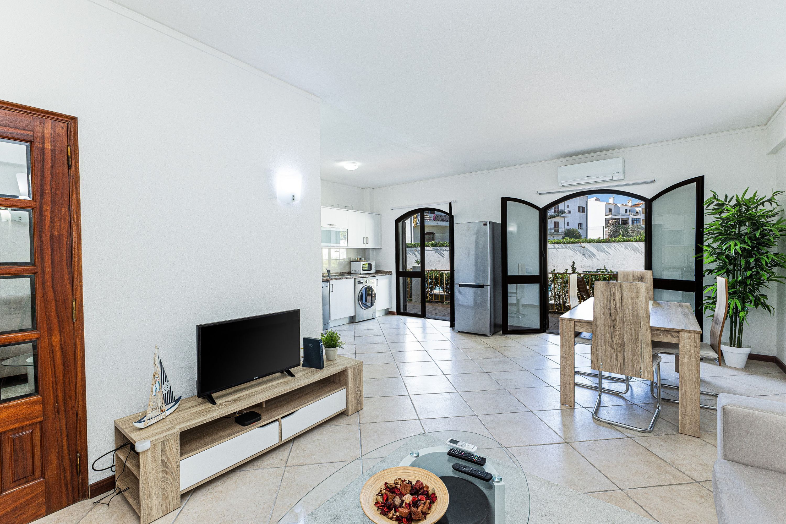 Una sala moderna con televisor, mueble de madera para entretenimiento, mesa de centro de vidrio y un rincón de comedor con silla y mesa.