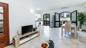 Uma sala de estar moderna com televisão, aparador de madeira, mesa de centro de vidro e um espaço de jantar com uma cadeira e mesa.