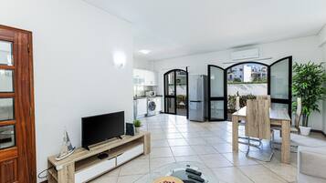 Una sala moderna con televisor, mueble de madera para entretenimiento, mesa de centro de vidrio y un rincón de comedor con silla y mesa.