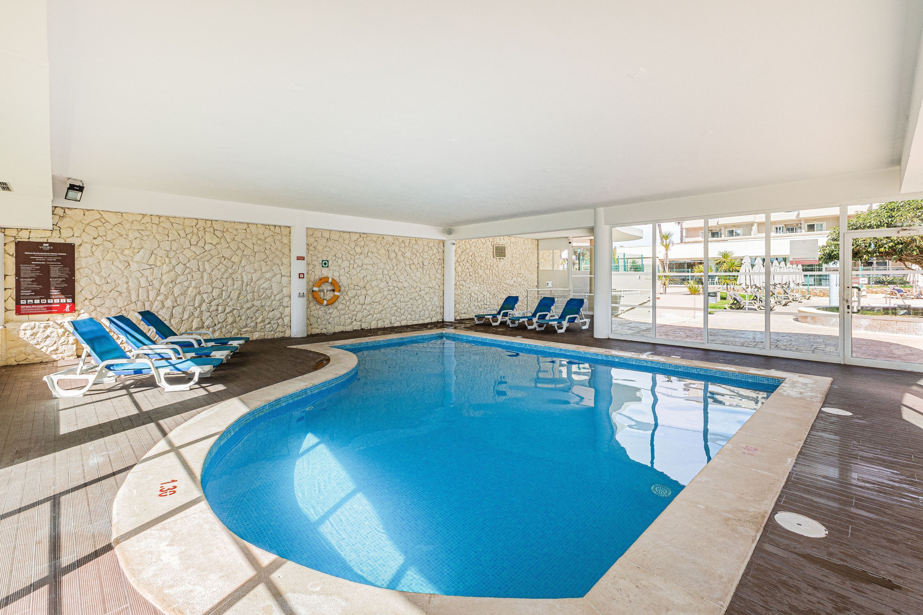 Una piscina interior con agua azul, rodeada de sillas de descanso y una pared de piedra.