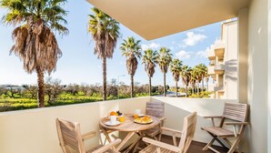 Ein Balkon mit Holztisch und Stühlen, ein Frühstücksbereich und Blick auf Palmen sowie ein Gebäude.
