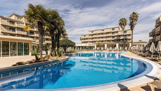 Ein Swimmingpool, umgeben von Palmen und einem mehrstöckigen Hotelgebäude.