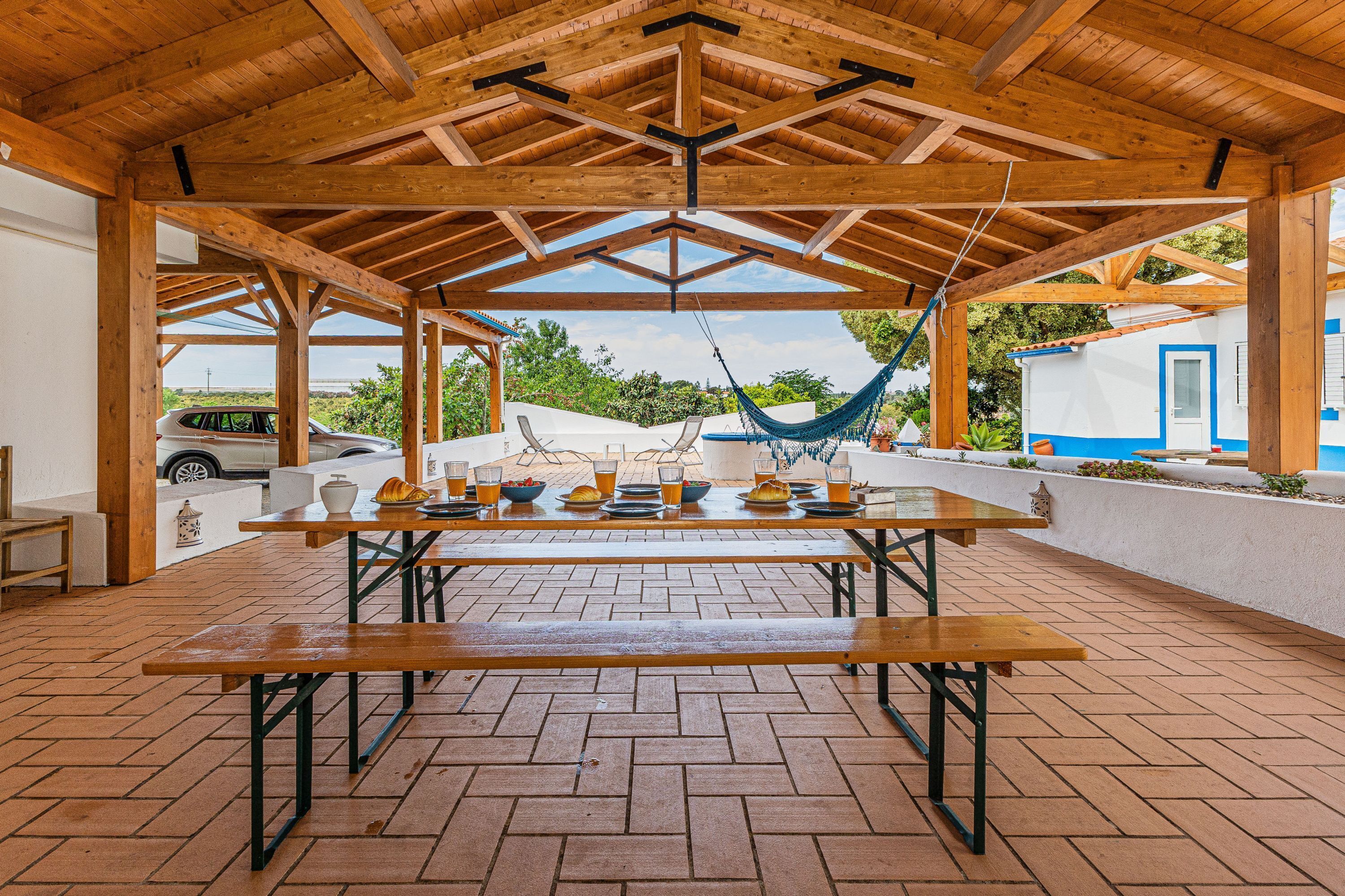 A covered patio with a wooden table set for dining, a hammock, and a view of a blue and white building.