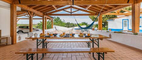 A covered patio with a wooden table set for dining, a hammock, and a view of a blue and white building.