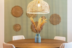 Ein runder Holztisch mit einer blauen Vase und getrockneten Pflanzen, umgeben von weißen Stühlen, unter einer gewebten Pendelleuchte, mit gewebten Wanddekorationen.