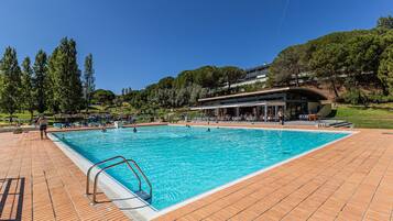 A swimming pool with a tiled deck, surrounded by trees and a building with a curved roof.