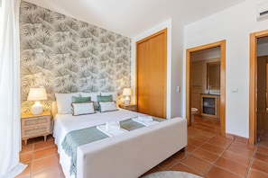 Ein Schlafzimmer mit Bett, Nachttischen, einer Wand mit Blattmuster, einer Holztür und einem durchgehenden Durchgang zu einem Badezimmer.