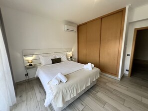 A neatly made bed with white linens and towels, a bedside lamp, a wooden wardrobe, and a wall-mounted air conditioner. - Praia da Rocha Pet Friendly By the Beach (Portimão)