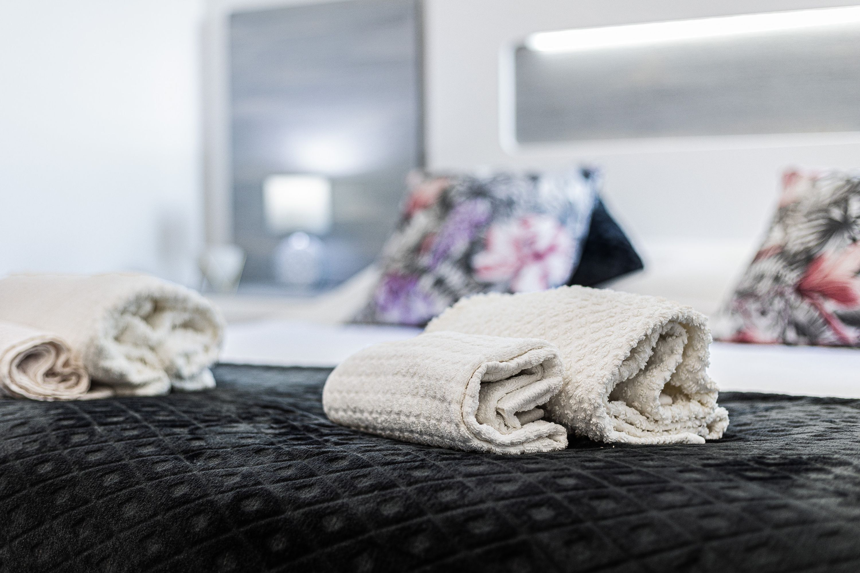 A neatly made bed with a rolled white towel and a folded white towel on a dark quilted bedspread.