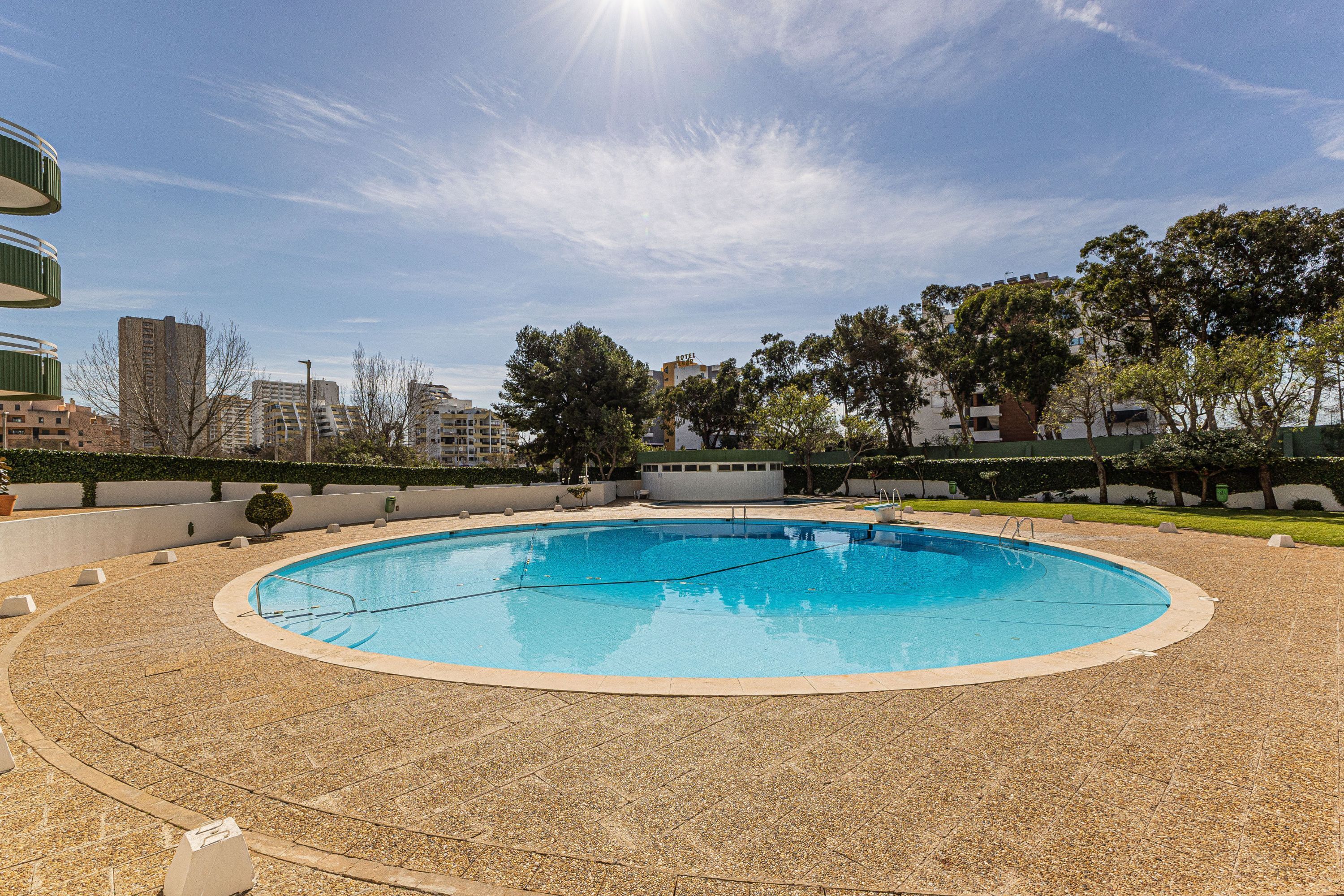 A swimming pool with a tiled deck, surrounded by trees and buildings.