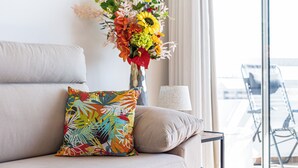 Un salon avec un canapé, un coussin coloré, un vase de fleurs, une lampe et une vue par la fenêtre.