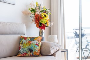 Ein Wohnzimmer mit einer Couch, einem bunten Kissen, einer Vase mit Blumen, einer Lampe und Blick durch ein Fenster.